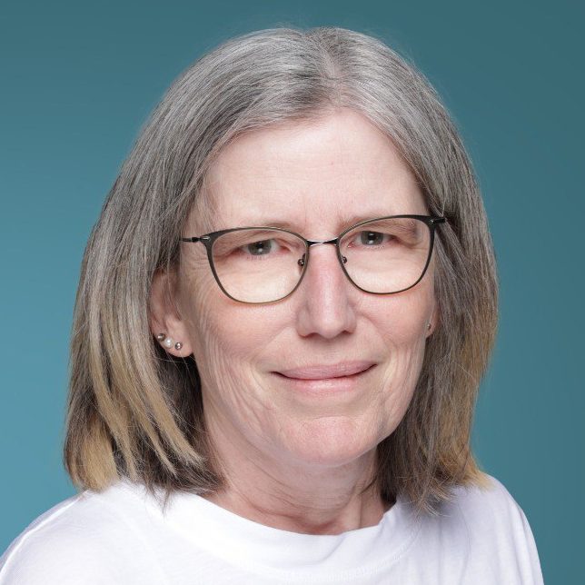 Porträt einer Frau mit Brille und mittellangen grauen Haaren, in einem hellen T-Shirt.