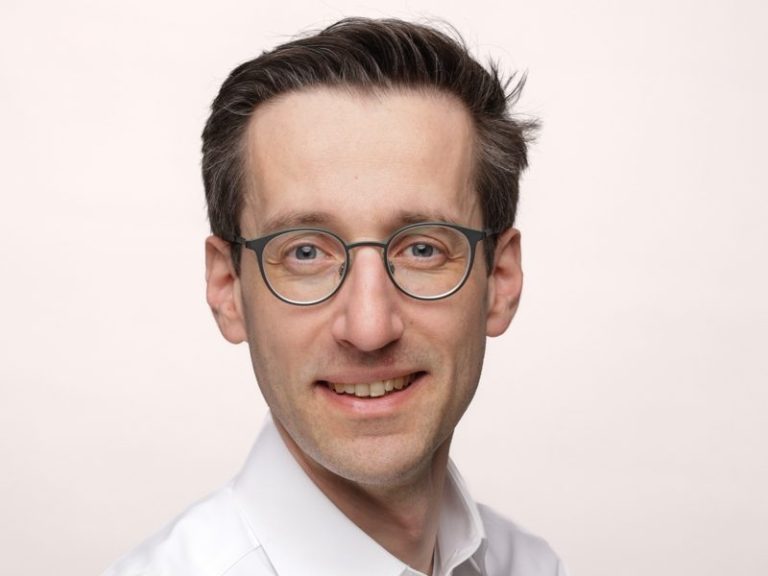 Portrait einer Person mit Brille, lächelnd, hellem Hintergrund.