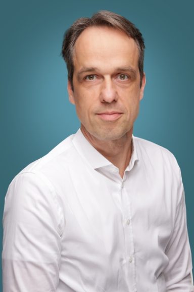 Mann mit kurzen Haaren in weißem Hemd vor einem einfarbigen Hintergrund.