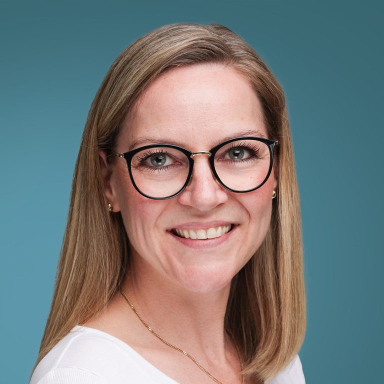 Frau mit blonden Haaren und Brille lächelt vor einem blauen Hintergrund.