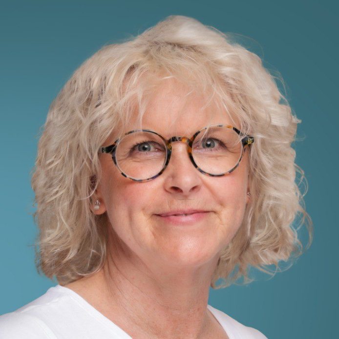 Porträt einer lächelnden Frau mit lockigem, blondem Haar und Brille, vor blauem Hintergrund.