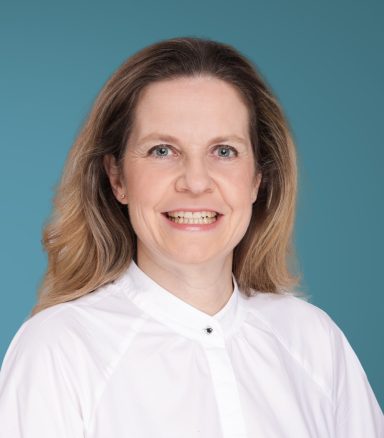 Porträt einer lächelnden Frau mit blonden Haaren vor einem blauen Hintergrund.