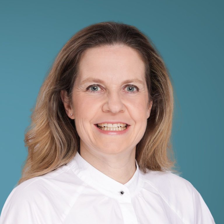 Praxisärztin Katrin Nückel, Porträt einer lächelnden Frau mit langen, blonden Haaren vor einem blauen Hintergrund.