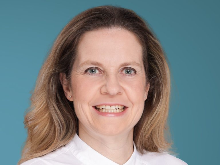 Frau mit blonden, lockigen Haaren und freundlichem Lächeln vor blauem Hintergrund.