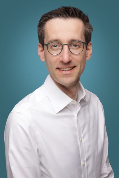 Porträt eines Mannes mit Brille und hellem Hemd vor einem blauen Hintergrund.