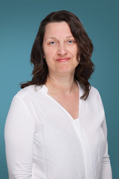 Frau mit langen, dunklen Haaren, trägt ein weißes Oberteil, lächelt freundlich.