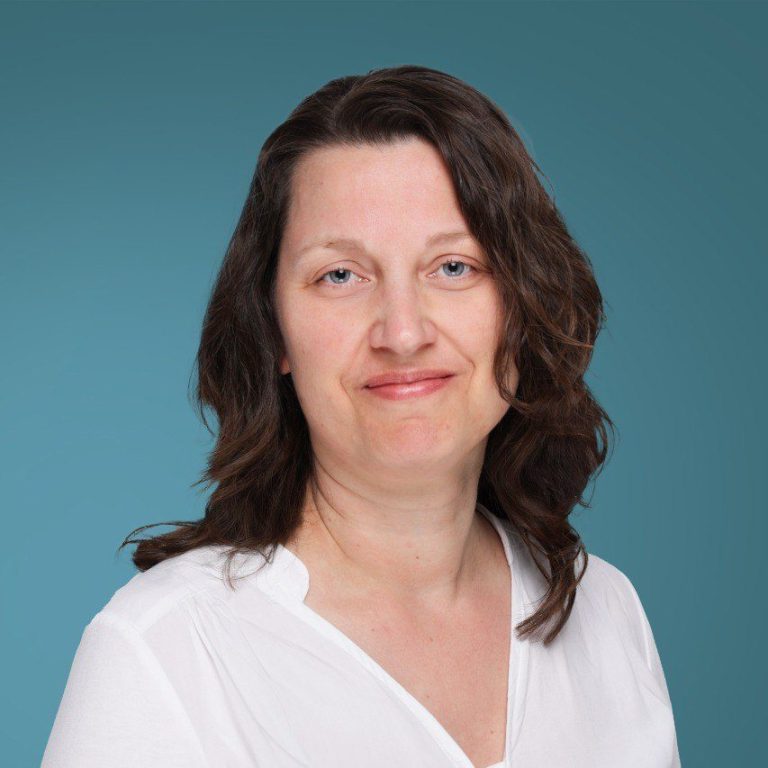 Frau mit braunen Haaren, lächelnd, trägt ein weißes Oberteil vor einem blauen Hintergrund.