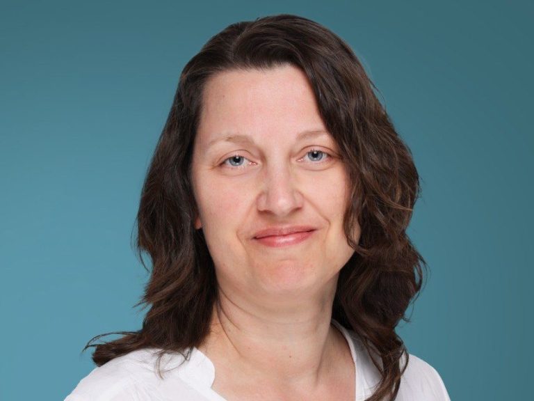 Frau mit lockigem, braunem Haar und lächelndem Gesicht vor einem blauen Hintergrund.