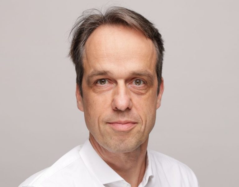 Porträt eines Mannes mit kurzen, grauen Haaren und einem weißen Hemd vor neutralem Hintergrund.