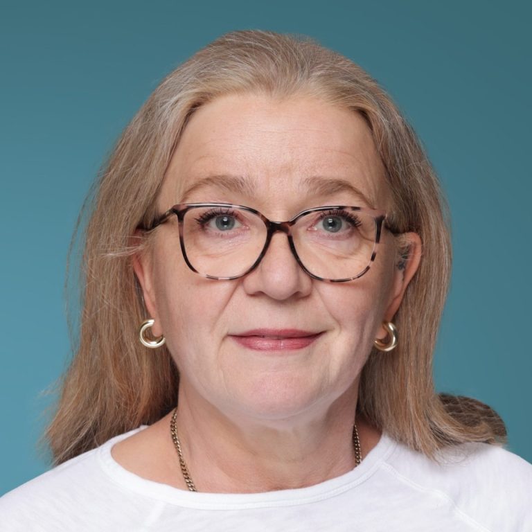 Frau mit Brille, blonden Haaren und einem freundlichen Ausdruck auf blauem Hintergrund.